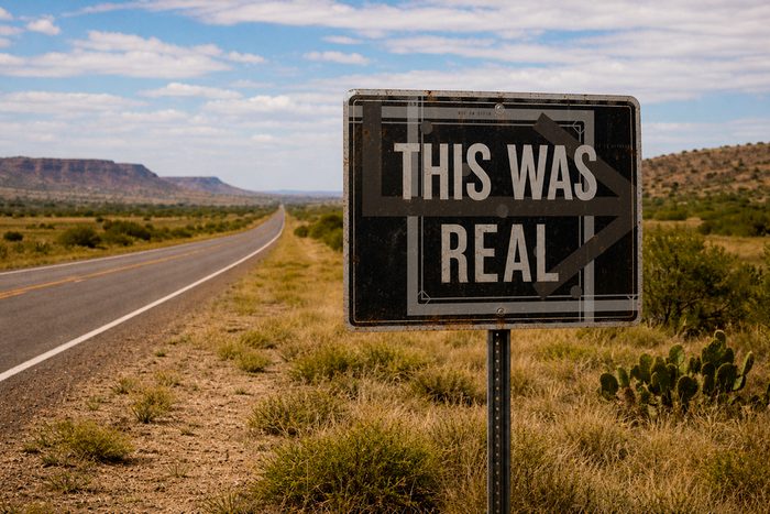 Dark sign reading THIS WAS REAL standing beside a desert highway under a bright sky