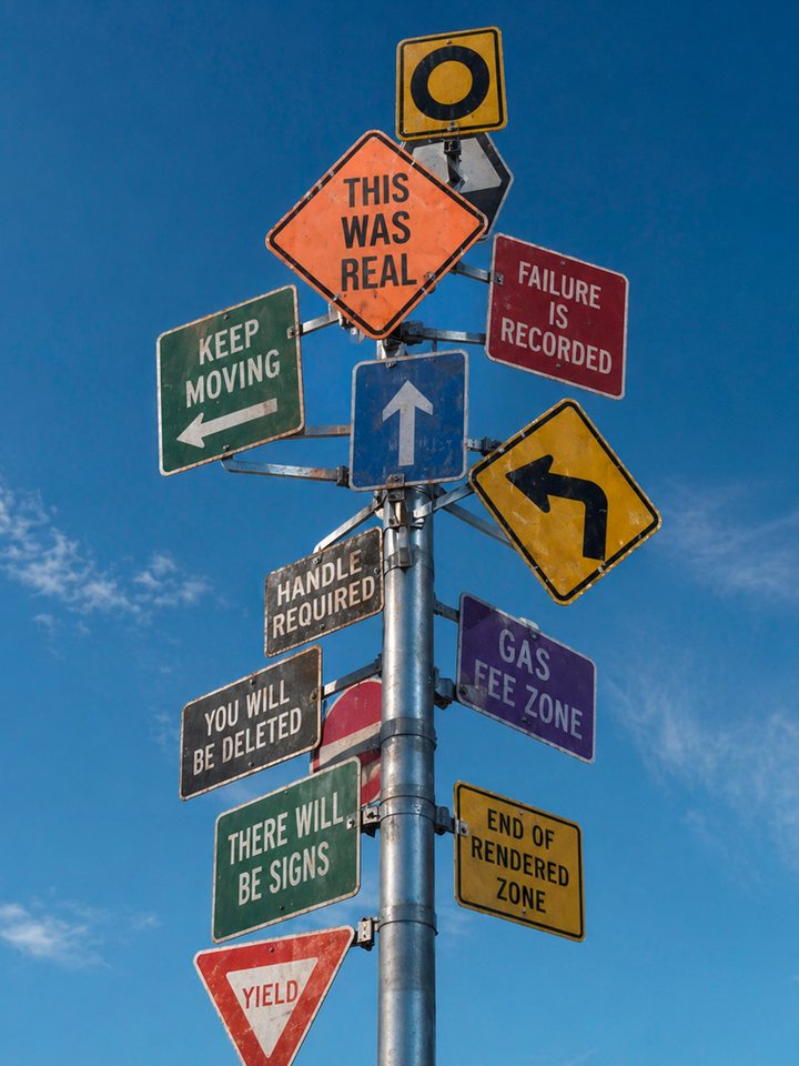 Cluster of signs crowded on a single post seen from below against deep blue sky, reading THIS WAS REAL, KEEP MOVING, YOU WILL BE DELETED and END OF RENDERED ZONE among others