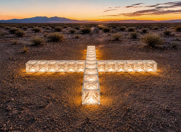 Cross of glass blocks lit from within at dusk, laid into the desert floor with mountains on the horizon